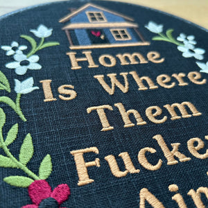 Black embroidered hoop art with a stitched house, flowers, and the funny quote ‘Home Is Where They Ain’t.’ Handmade humorous wall decor
