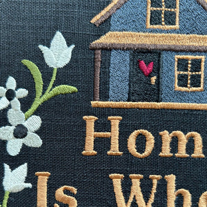 Embroidered design of a house with flowers and 'Home is Where' text on a dark background