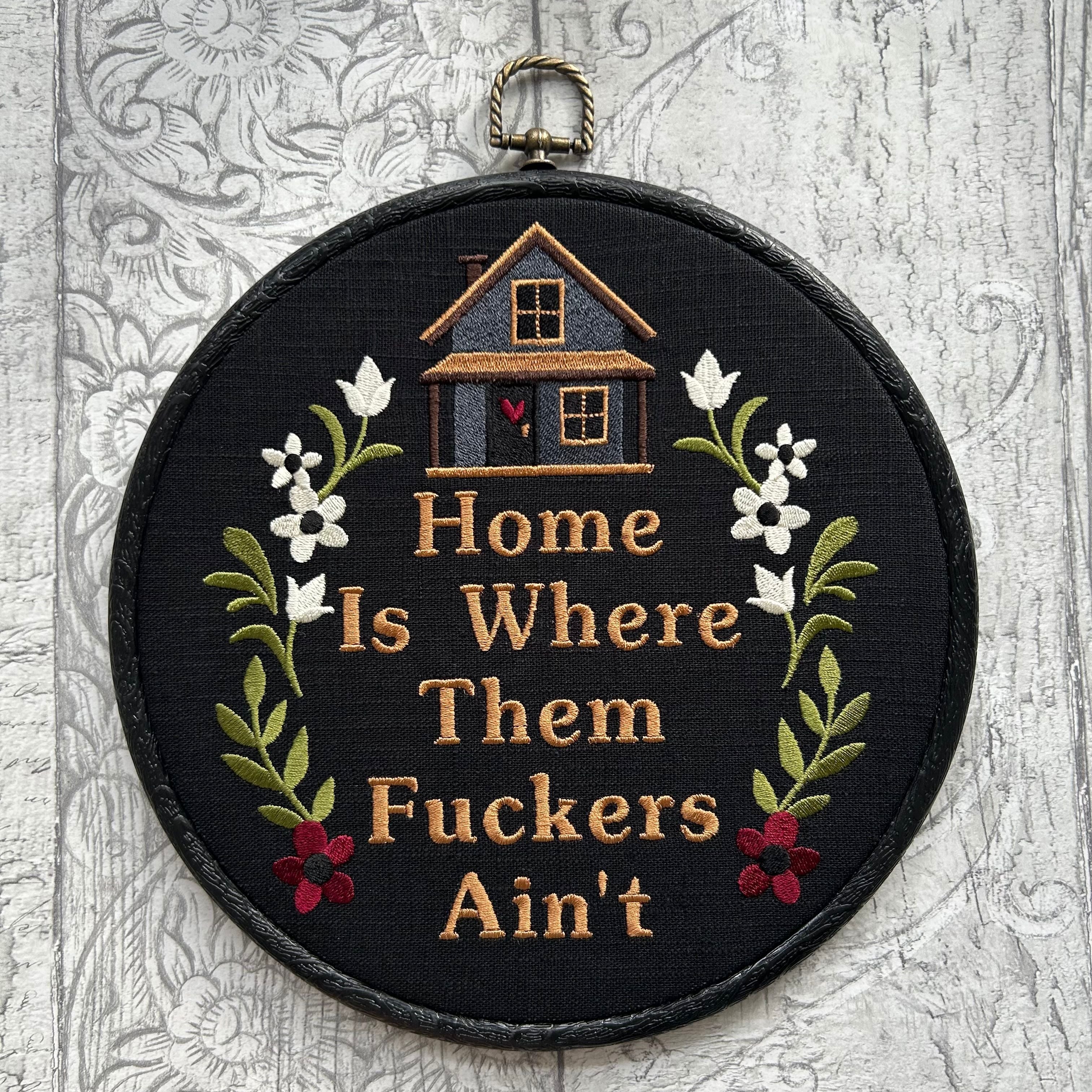 Black embroidered hoop art featuring a small stitched house with floral accents and the humorous phrase ‘Home Is Where Them [Expletive] Ain’t.’ Handmade funny home decor with rustic charm.