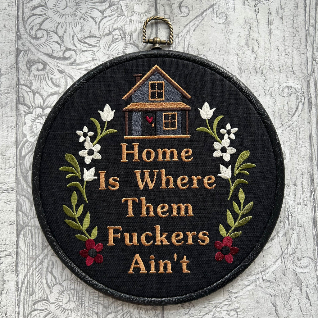 Black embroidered hoop art featuring a small stitched house with floral accents and the humorous phrase ‘Home Is Where Them [Expletive] Ain’t.’ Handmade funny home decor with rustic charm.