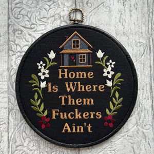 Black embroidered hoop art featuring a small stitched house with floral accents and the humorous phrase ‘Home Is Where Them [Expletive] Ain’t.’ Handmade funny home decor with rustic charm.