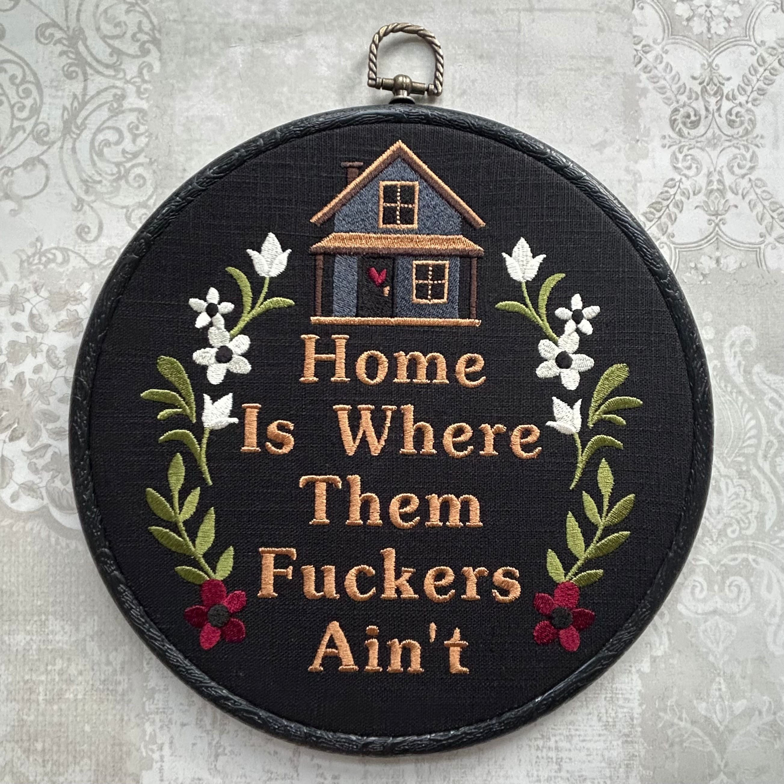 Embroidery hoop with text 'Home Is Where Them Fuckers Ain't' and house illustration on a floral background.