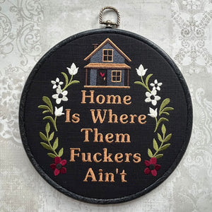 Embroidery hoop with text 'Home Is Where Them Fuckers Ain't' and house illustration on a floral background.